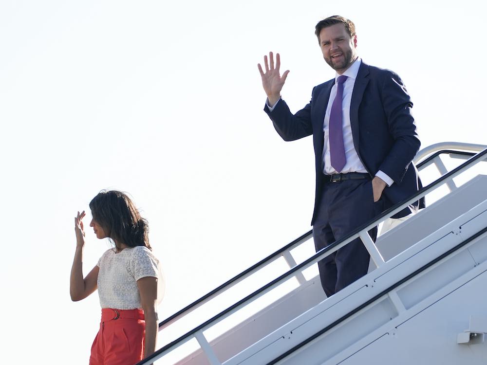 J.D. Vance descends a white flight of airplane stairs. He pauses mid-step to wave at the camera. He is wearing a navy suit over a white shirt and purple tie. He has short brown hair, a beard and tanned light skin. Ahead of him is his spouse, Usha Vance, a woman with medium skin, long dark hair, a white top and a red skirt. Behind them, the sky is bright white.