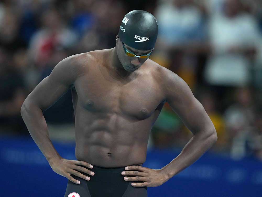 Josh Liendo is a Black man standing with his hands on his hips. He wears a black swim cap, goggles and black swimming bottoms. Behind him is an audience in soft focus.