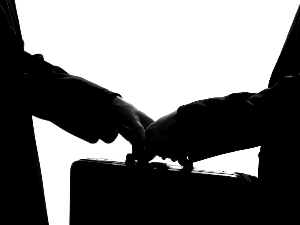 A contrasty black and white image shows the arms of two suit-clad people, each with one hand on the handle of a suitcase between them.