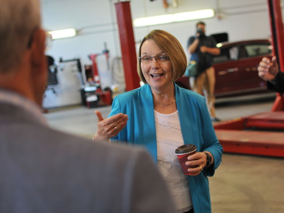 A woman with light skin and dark blond hair in a bob is smiling as she speaks with a person with their back to the camera. She is wearing a blue blouse over a white top and holds a takeout coffee cup in one hand.