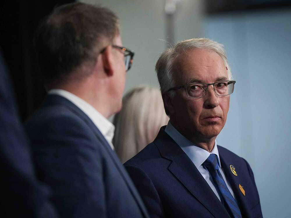 John Rustad, right, is a white man with short white hair and glasses. He frowns at Kevin Falcon to his left, a white man with short brown hair, glasses and his back to the camera.