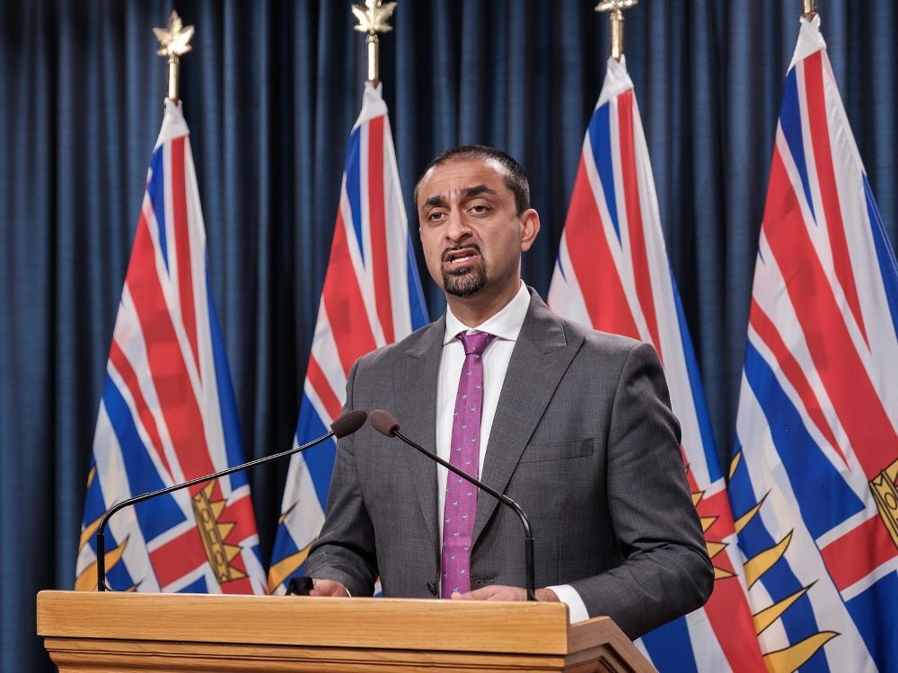 A person with medium-toned skin and wearing a suit stands at a podium with B.C. flags behind him.