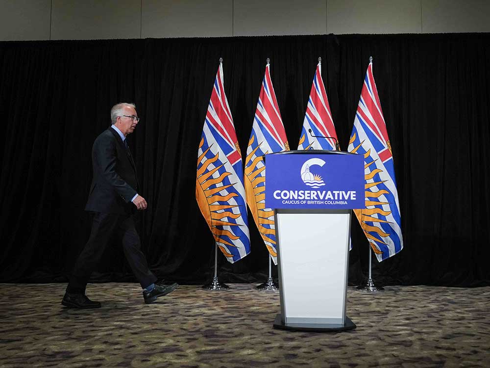 John Rustad walks from the left of the screen in a darkened room towards a blue podium that reads 'Conservative Caucus of British Columbia.' Behind the podium are four B.C. flags in blue, white, red and yellow.