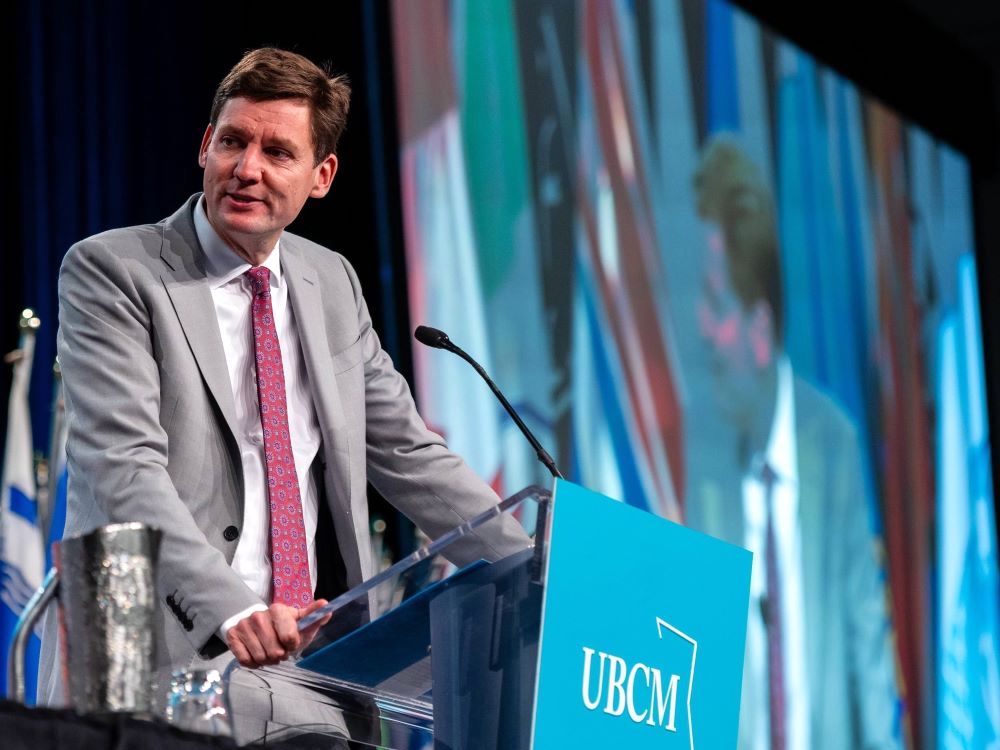David Eby is a tall white man with short brown hair. He wears a light grey suit and red patterned tie over a white button-down shirt. He leans against a podium with a turquoise 'UBCM' sign on it and looks towards the left of the frame. Behind him is a row of flags and a large screen broadcasting an image of Eby at the podium.