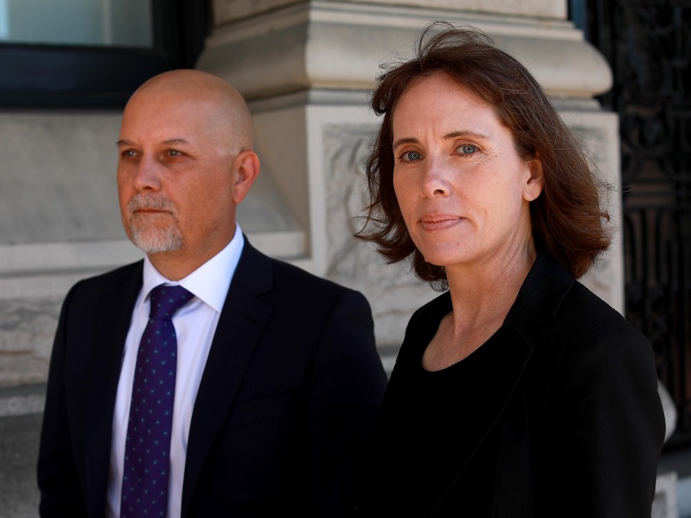 On the left, a bald man with light skin and a grey moustache and goatee, wearing a dark jacket over a white shirt and blue tie. On the right, a woman with light skin and shoulder-length brown hair, wearing black and looking at the camera with a slight smile.