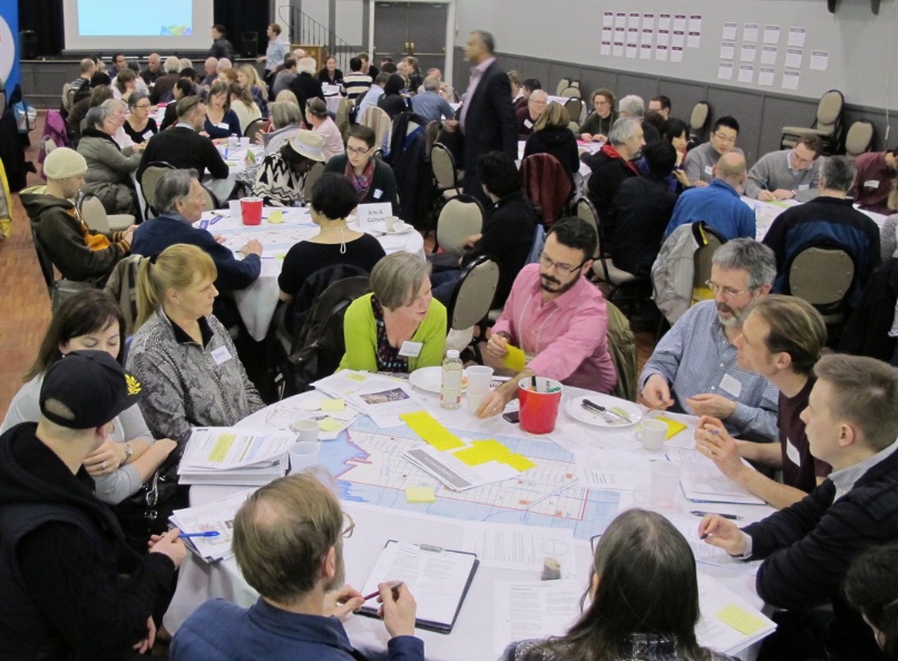A room full of people sitting at round tables with diagrams spread over them.