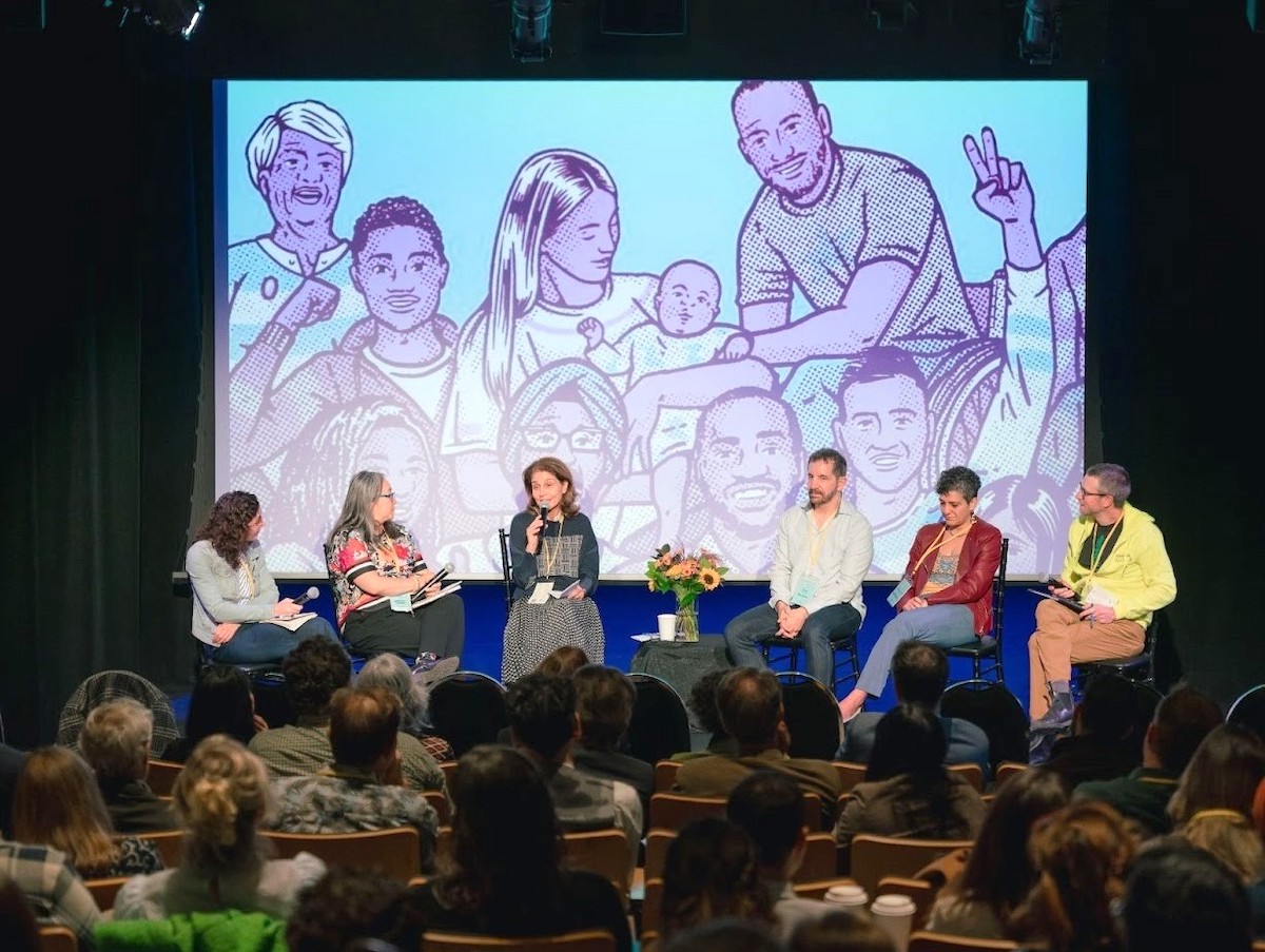 A panel of six people sit on a stage in front of an audience. One woman on the panel speaks into a microphone. Behind them is a large screen with a purple-hued illustration of a diverse group of people.