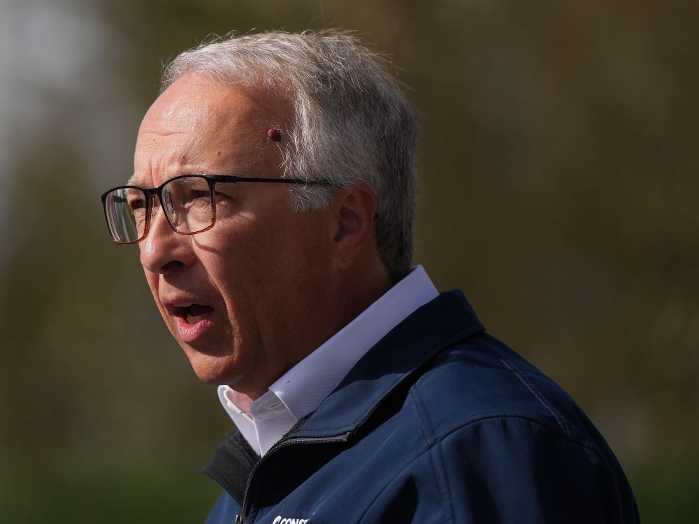 Profile of a 60-ish man with light skin tone, short grey hair and glasses, wearing a navy blue jacket over a white collared shirt. His mouth is open as if in mid-speech.