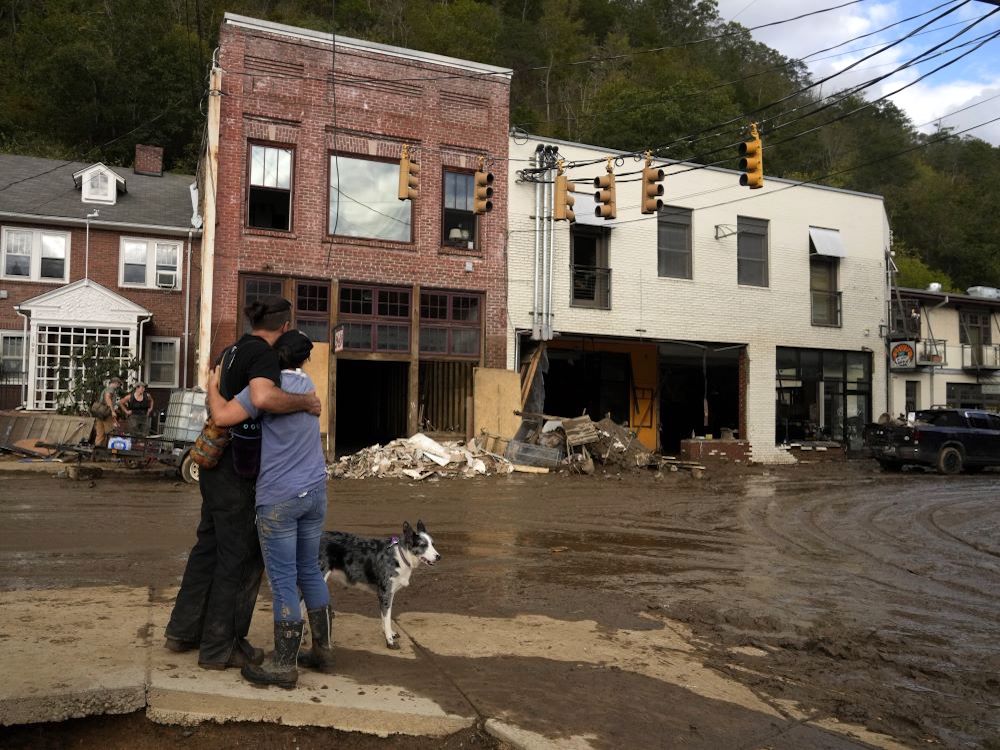 Two people with their back to the camera embrace while standing in a muddy street, looking out over hurricane damage in the town of Marshall, North Carolina.