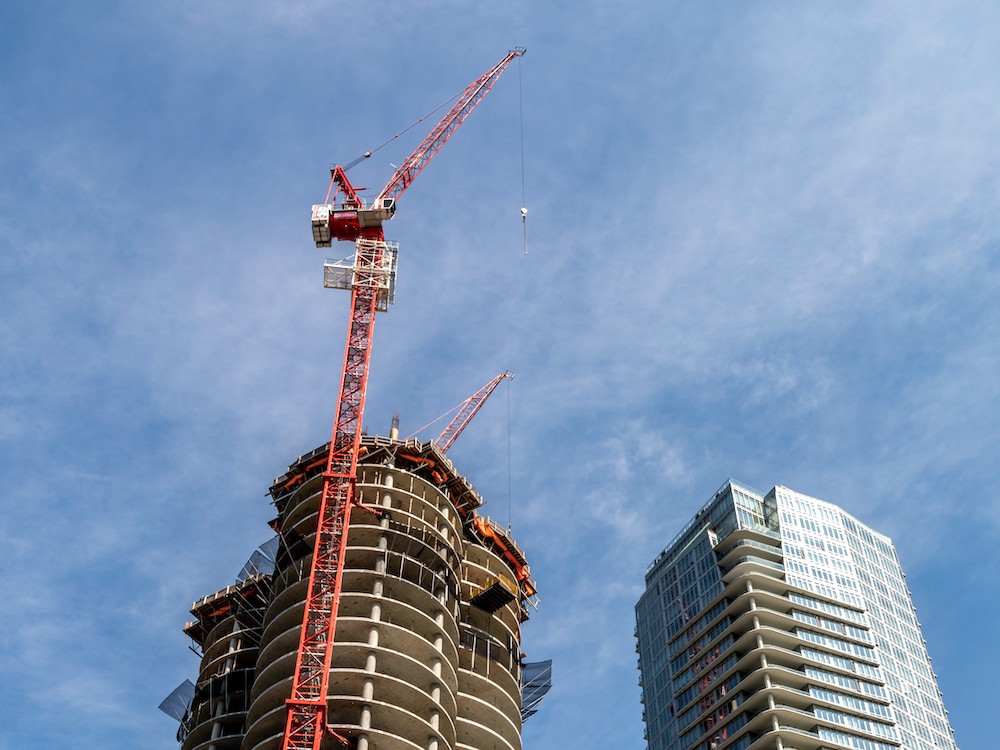Viewed from below, a highrise under construction and a taller red crane rise into a blue sky.