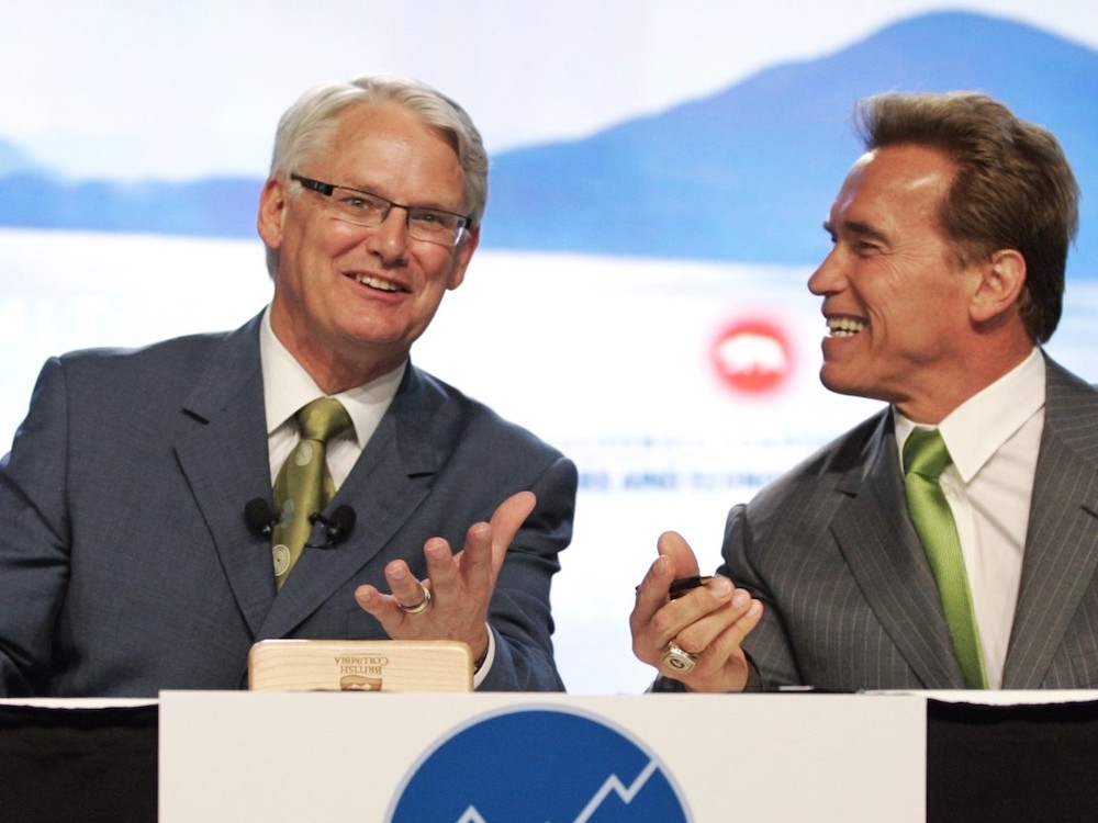 Gordon Campbell, a man with light skin, grey hair and glasses sits behind a table next to Arnold Schwarzenegger, a man with light skin and brown hair. Both men are smiling and gesturing in conversation.