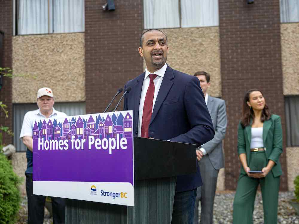 Ravi Kahlon has medium skin, short dark hair and a short beard. He wears a navy suit over a white button-down shirt and red tie. He stands at a podium with an affixed purple sign that reads 'Homes for people.' Three other people stand behind him; behind them all is a brown brick apartment building.