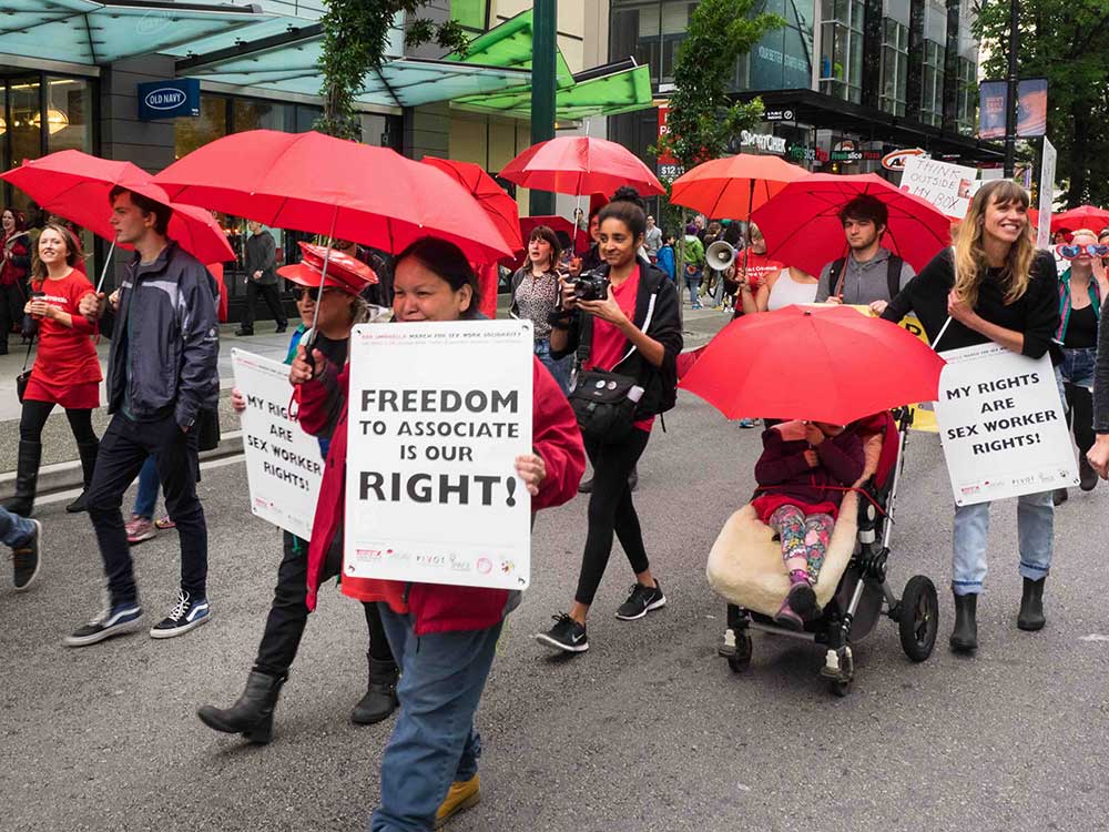 People march down a downtown Vancouver street holding red umbrellas on a grey day. Two people in the foreground hold white placards that read &ldquo;My rights are sex worker rights!&rdquo; and &ldquo;Freedom to associate is our right!&rdquo;