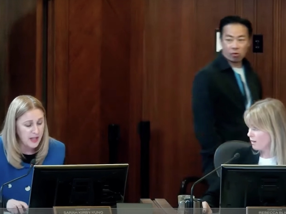 Rebecca Bligh, at the right of the frame, looks incredulously at Sarah Kirby-Yung, at left, while seated inside Vancouver council chambers. Both women have blond hair and light skin and are wearing office attire. Behind them is extensive wood panelling. In the background to the right, Mayor Ken Sim walks by and looks on. He is a Chinese man with short black hair and medium skin; his hands are in his pockets.