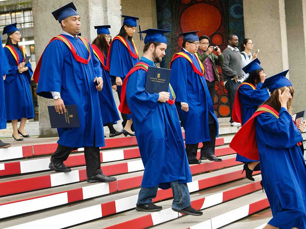 A large group of people descend a red and white stairway in blue graduation caps and gowns with red and yellow sashes.
