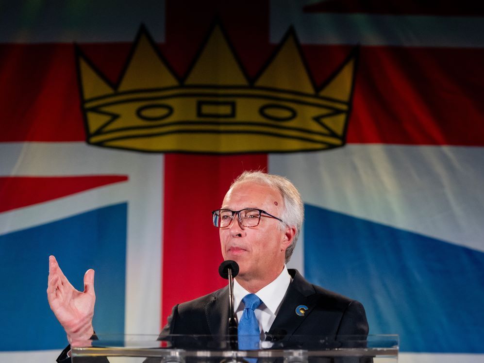 A grey-haired white man wearing glasses gestures at a podium, with a banner showing a crown on it behind him and above his head — a part of the image on the B.C. flag.