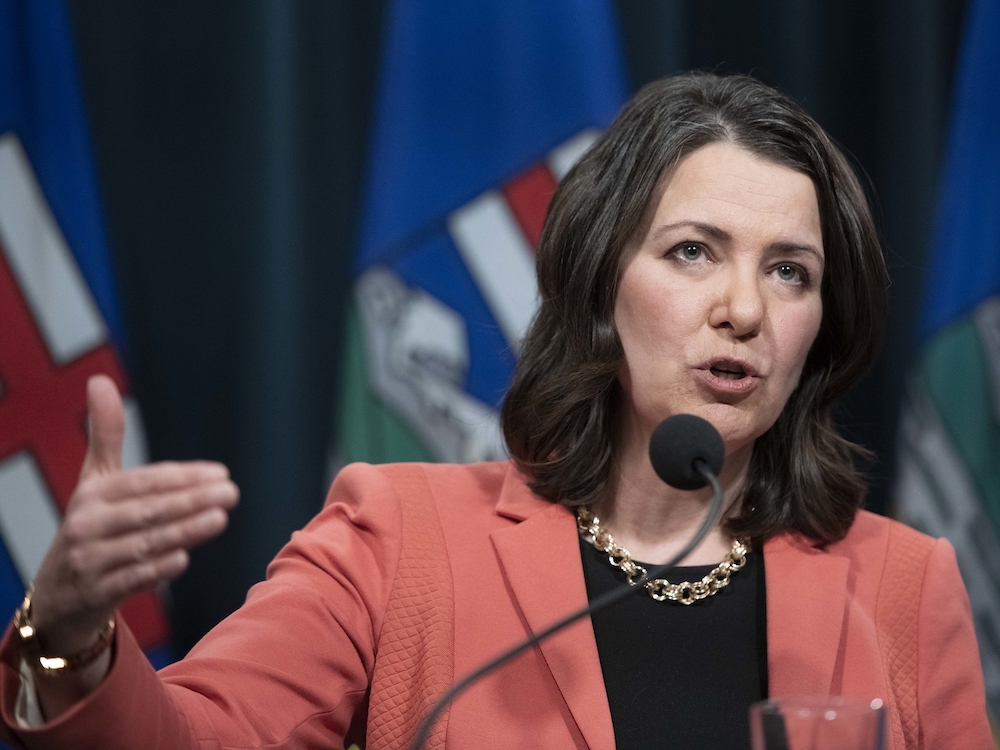 A 53-year-old light-skinned woman with shoulder-length dark brown hair gestures while standing at a podium. She wears a black shirt, salmon blazer and chunky gold necklace.