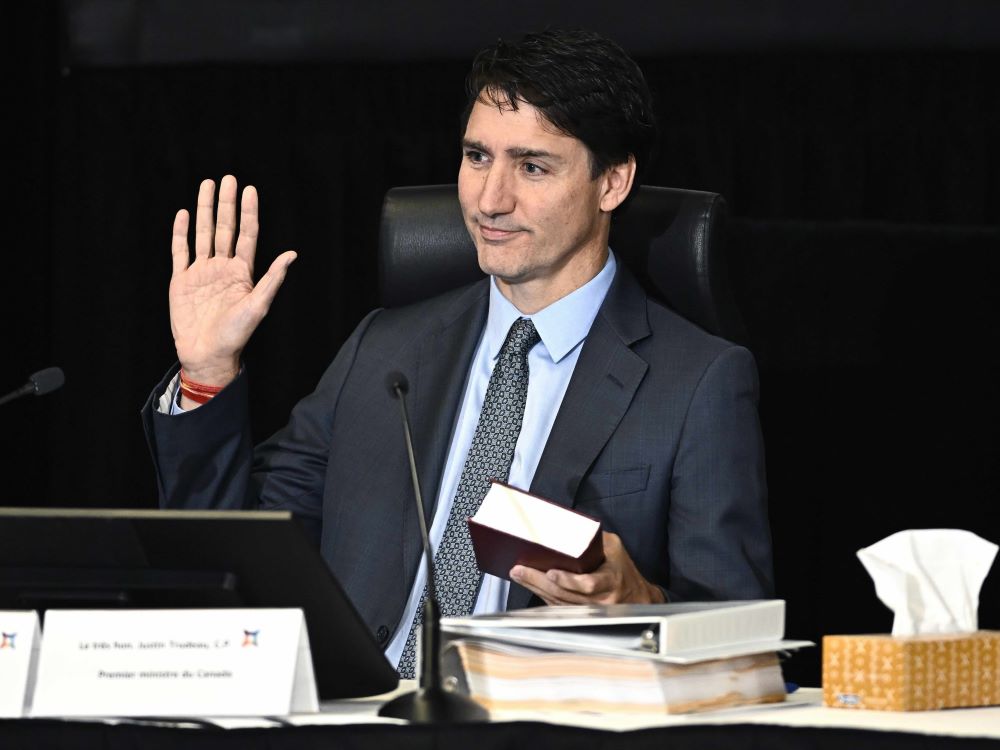 Justin Trudeau has short dark wavy hair, light skin and blue eyes. He is raising his right hand while holding a thick red book in the other. He wears a dark blue suit over a light blue button-down and patterned blue tie. In the foreground is a black tabletop microphone and a stack of binders to the right of the frame.