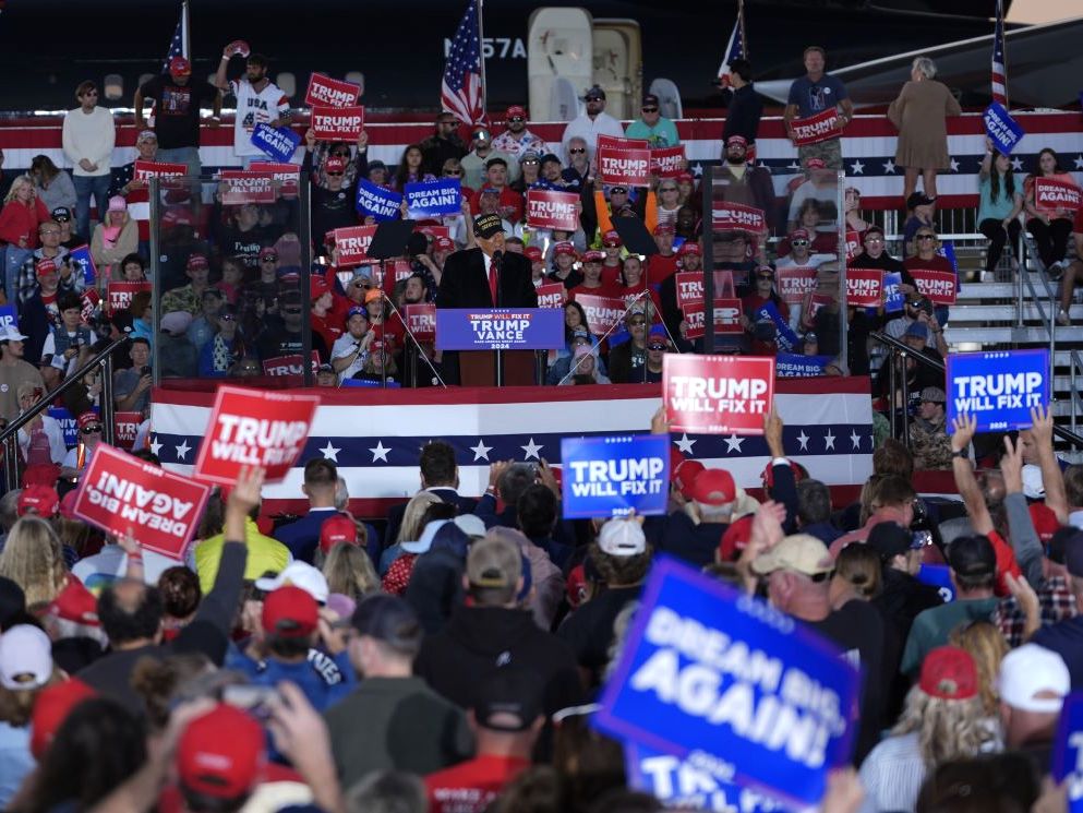 A hall full of raucous rally goers waving blue, white and red signs, some saying 'Trump Will Fix It,' with Donald Trump a small figure in the middle.