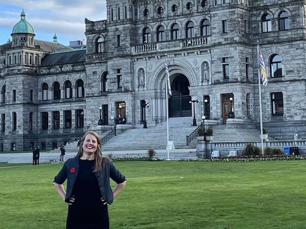 Christine Boyle has shoulder-length blond hair and light skin. She&rsquo;s smiling brightly at the camera and standing with her hands on her hips on the front lawn of the B.C. parliament buildings in Victoria; the buildings, grey with domed green copper roofs, are in the background. Boyle wears a dark denim jacket, a black dress and red sneakers.