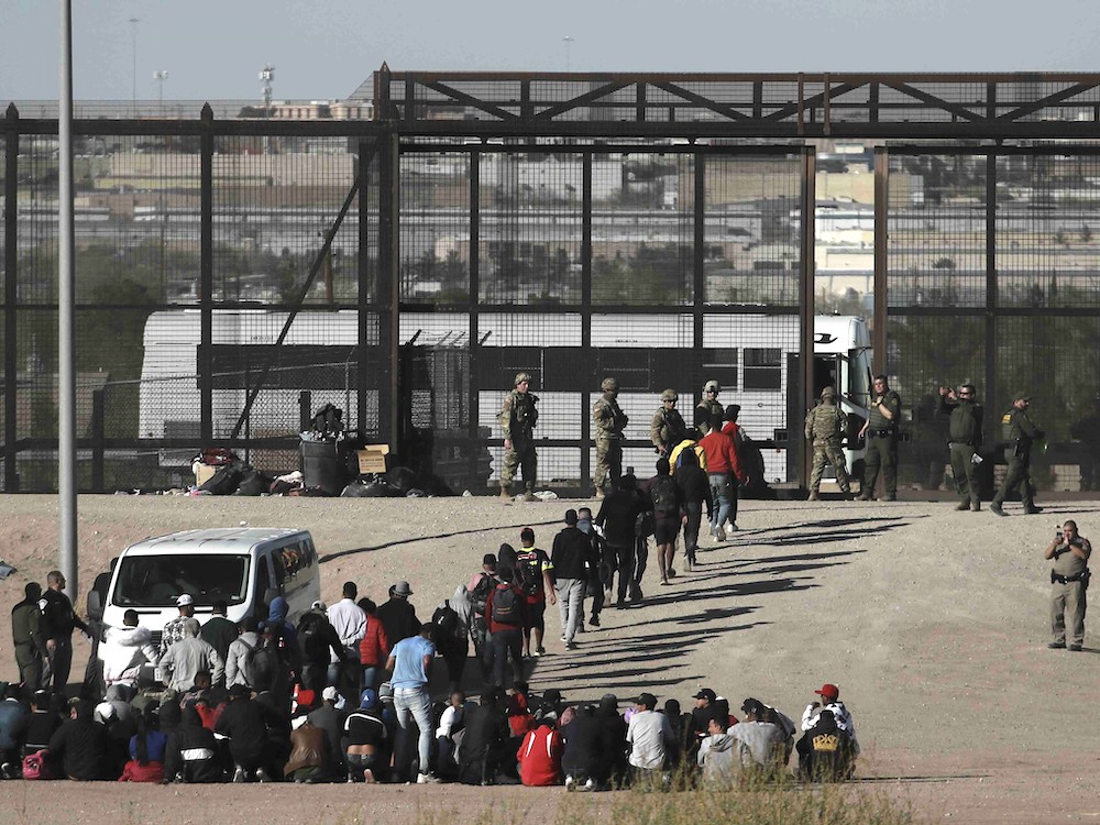 Crowds of people line up outside before a tall set of metal gates to board a white bus guarded by people in military fatigues.