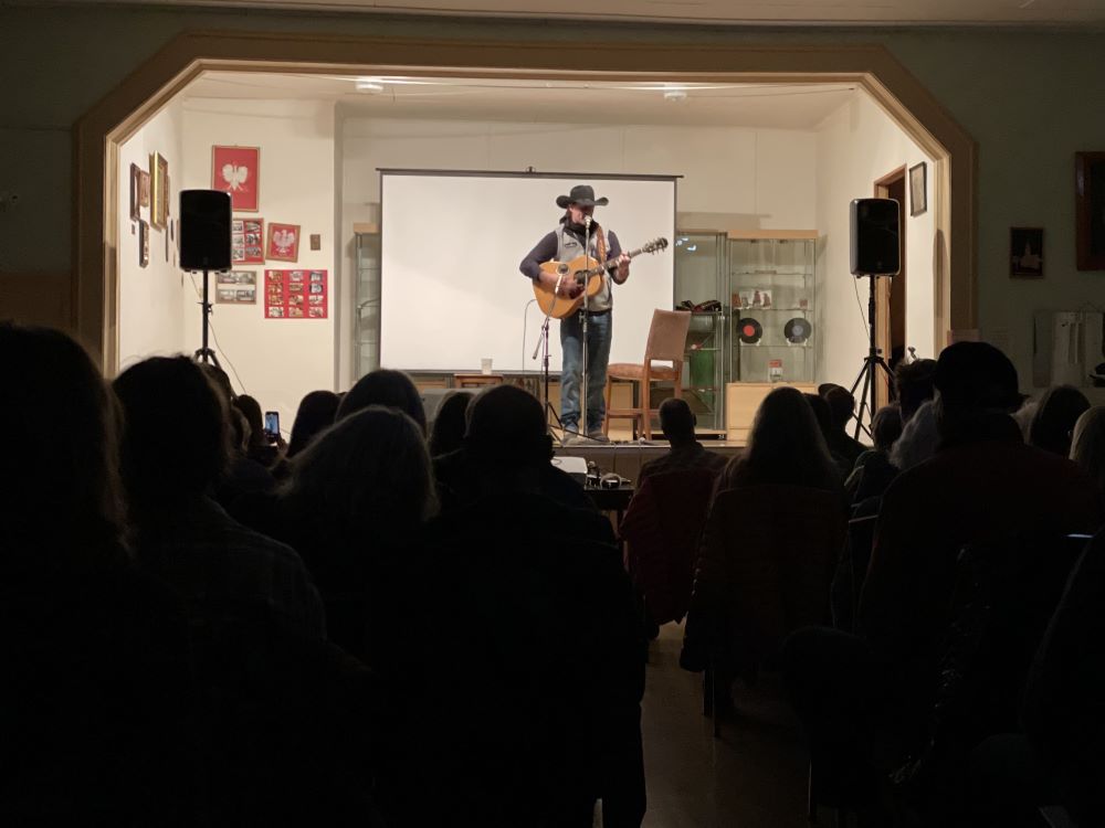 A man in a cowboy hat, country singer Corb Lund, plays a guitar and sings on stage in front of an audience.