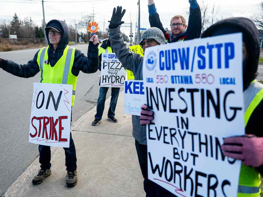 A half-dozen people stand by a roadway holding signs that say 'On strike.'