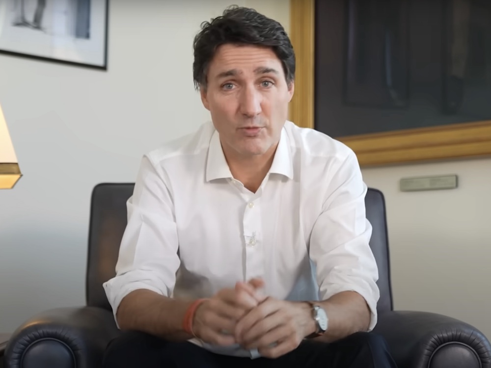 Justin Trudeau has wavy brown hair, a white button-down shirt and dark grey trousers. He is seated on a brown leather armchair and a beige lamp is to the left of the frame. He is pausing mid-speech.