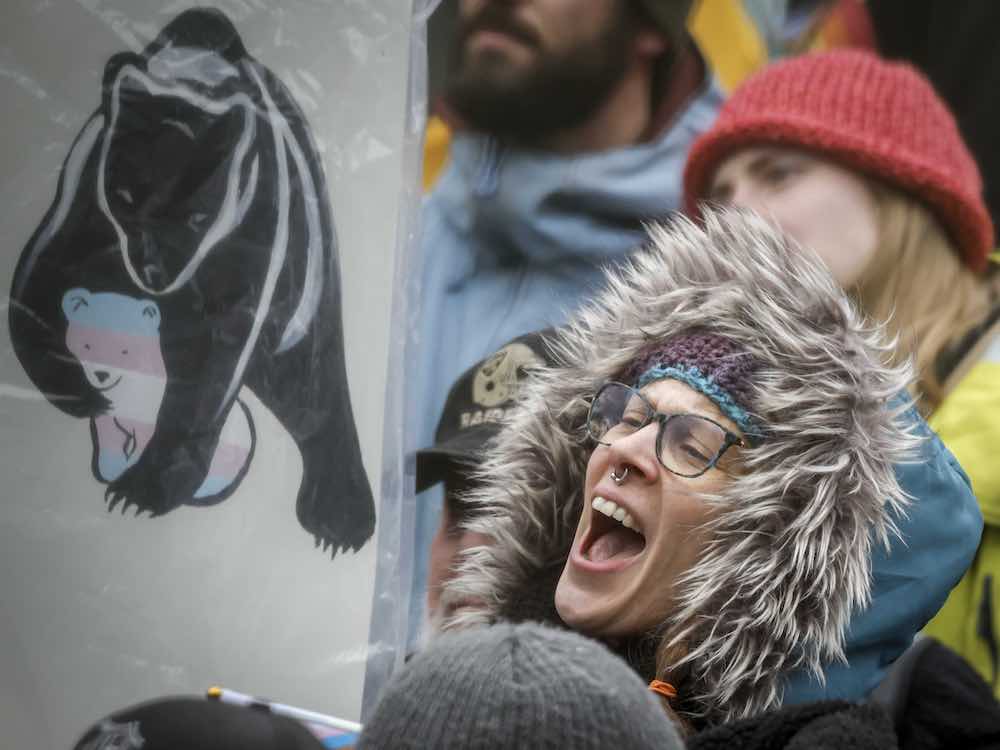 A person in glasses with a septum piercing stands in a crowd at a rally, shouting. They are wearing a turquoise jacket with a fur-lined hood that frames their face. To the left of the frame is a sign featuring an illustration of a black bear protecting a smaller bear coloured with the light blue, white and pink stripes of the transgender flag.