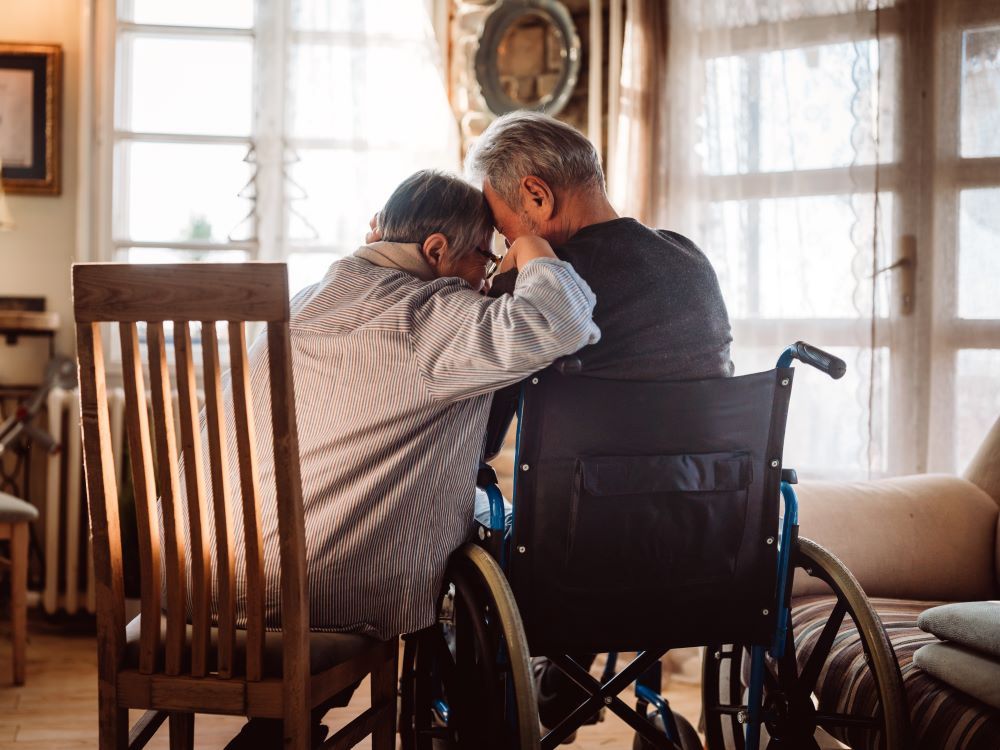 A couple of older people sitting together in a room as light pours in through big windows in front of them. One person is in a wooden chair and the other is sitting in a wheelchair.