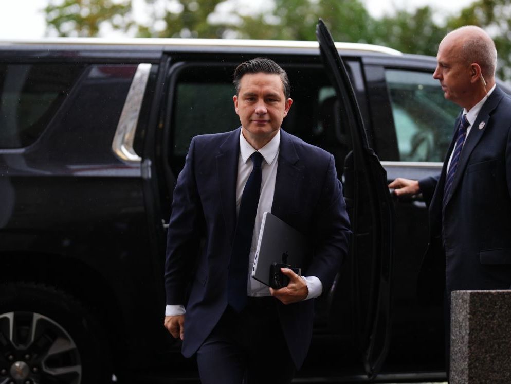 Pierre Poilievre, a man with light skin and dark slicked-back hair wearing a navy suit and tie, exits the back of a black SUV as a security guard holds the door open.