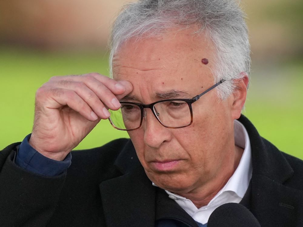 A middle-aged white man with grey hair and glasses looks contemplative and touches his brow.