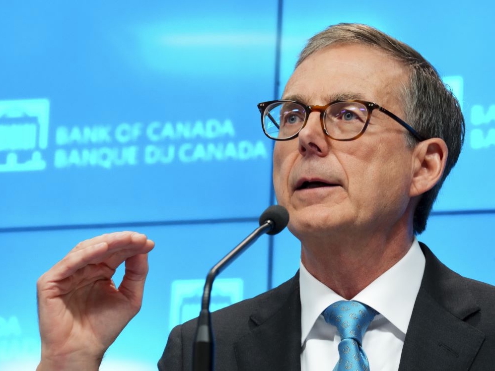 A middle-aged white man with grey hair and glasses gestures at a podium with the words 'Bank of Canada' behind him on a blue wall.