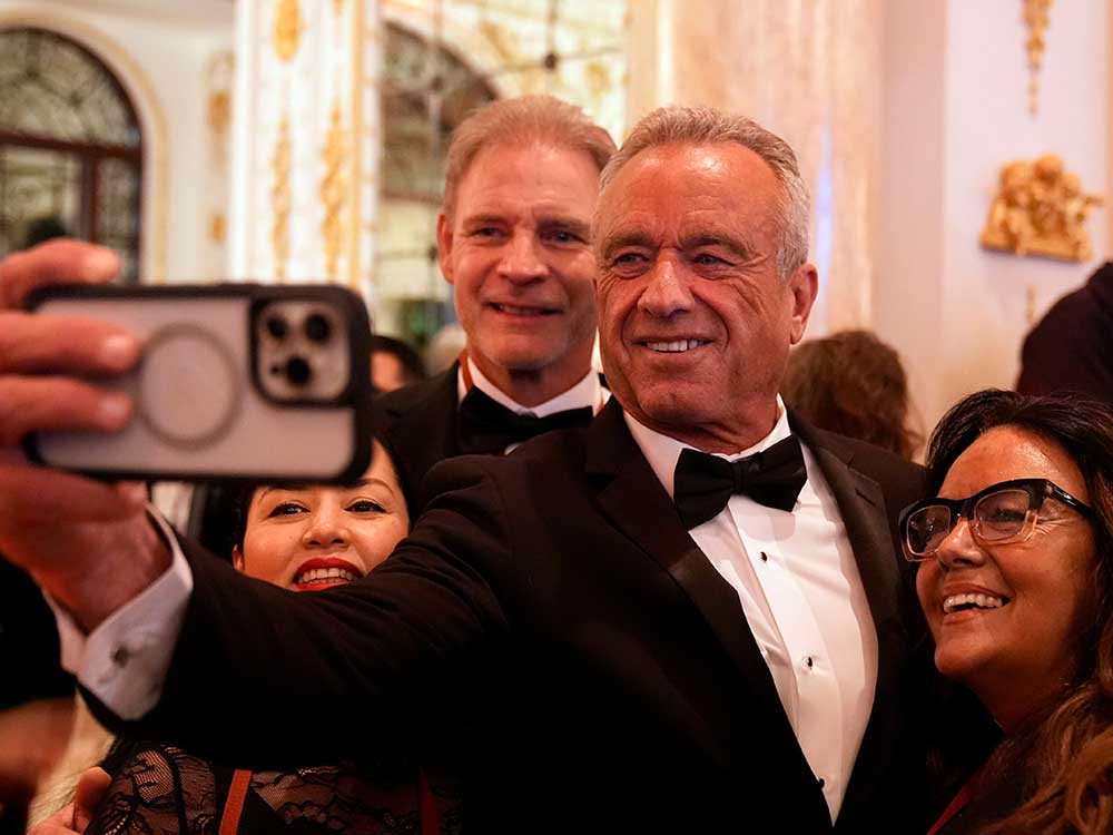 Robert F. Kennedy Jr. has short grey hair and tanned skin. He is wearing a tuxedo and has an arm outstretched, holding a smartphone, to take a selfie with two women and one man, all in black tie formalwear.