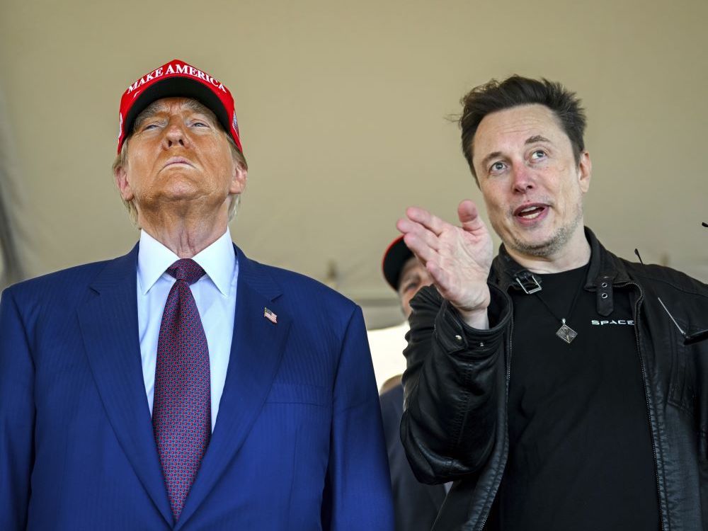 A 78-year-old man apparently wearing orange makeup, in a blue suit and a red MAGA hat, scowls as he stands beside a 50-ish light-skinned man wearing a black T-shirt and leather jacket.