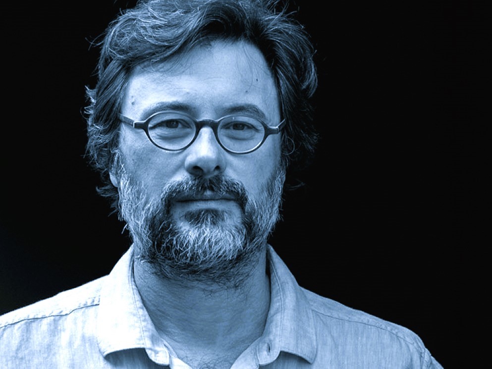 Headshot of a light-skinned man with wavy salt and pepper hair and beard and glasses, looking directly into the camera. The image is black and white with a bluish tint.