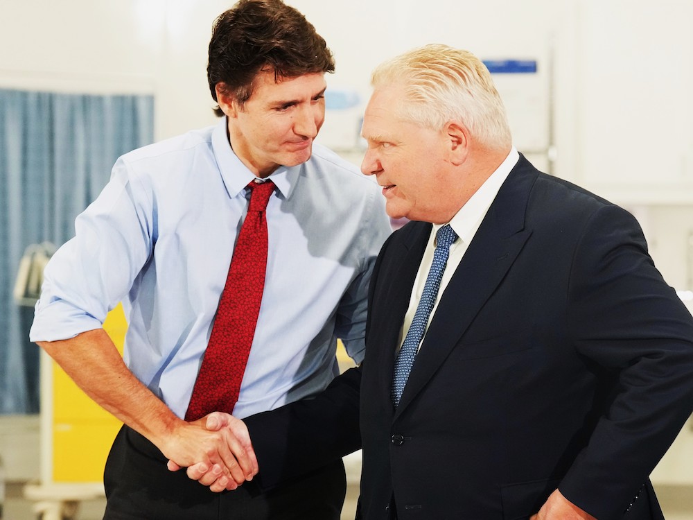 Justin Trudeau, wearing a light blue dress shirt with his sleeves rolled up and a red tie, and Doug Ford, wearing a black suit jacket over a white shirt and blue tie, in a firm-looking handshake.