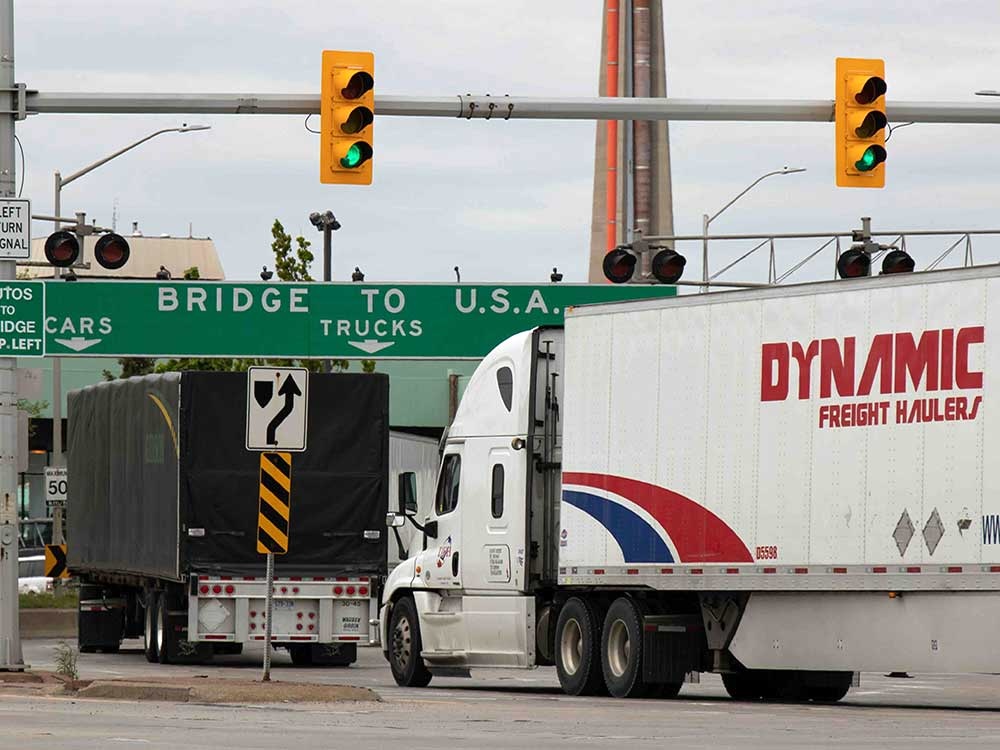 A large white transport truck is emblazoned with red text that reads 'Dynamic Freight Hauler.' It drives behind a black transport truck under a green sign that says 'Bridge to U.S.A.'