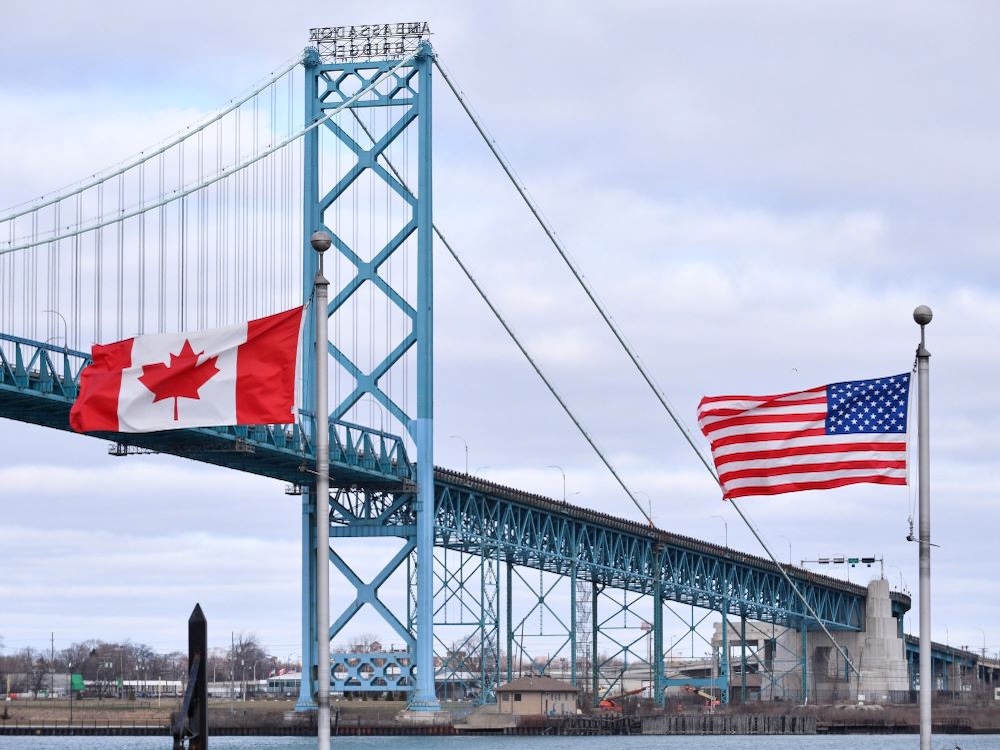 Canadian and American flags fly in front of a silver-grey suspension bridge.