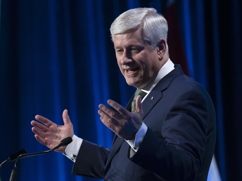 A 65-year-old light-skinned man with grey hair, wearing a blue suit, gestures as he stands at a podium.