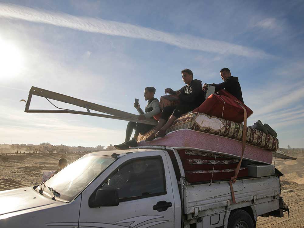 Three young men sit atop a stack of mattresses and other household items that are strapped to the bed of a white pickup truck. The man to the right is looking at the camera, smiling. They are wearing casual streetwear and seated against a blue sky. Behind them is a dry landscape with low buildings. A child stands outside the passenger seat of the truck.