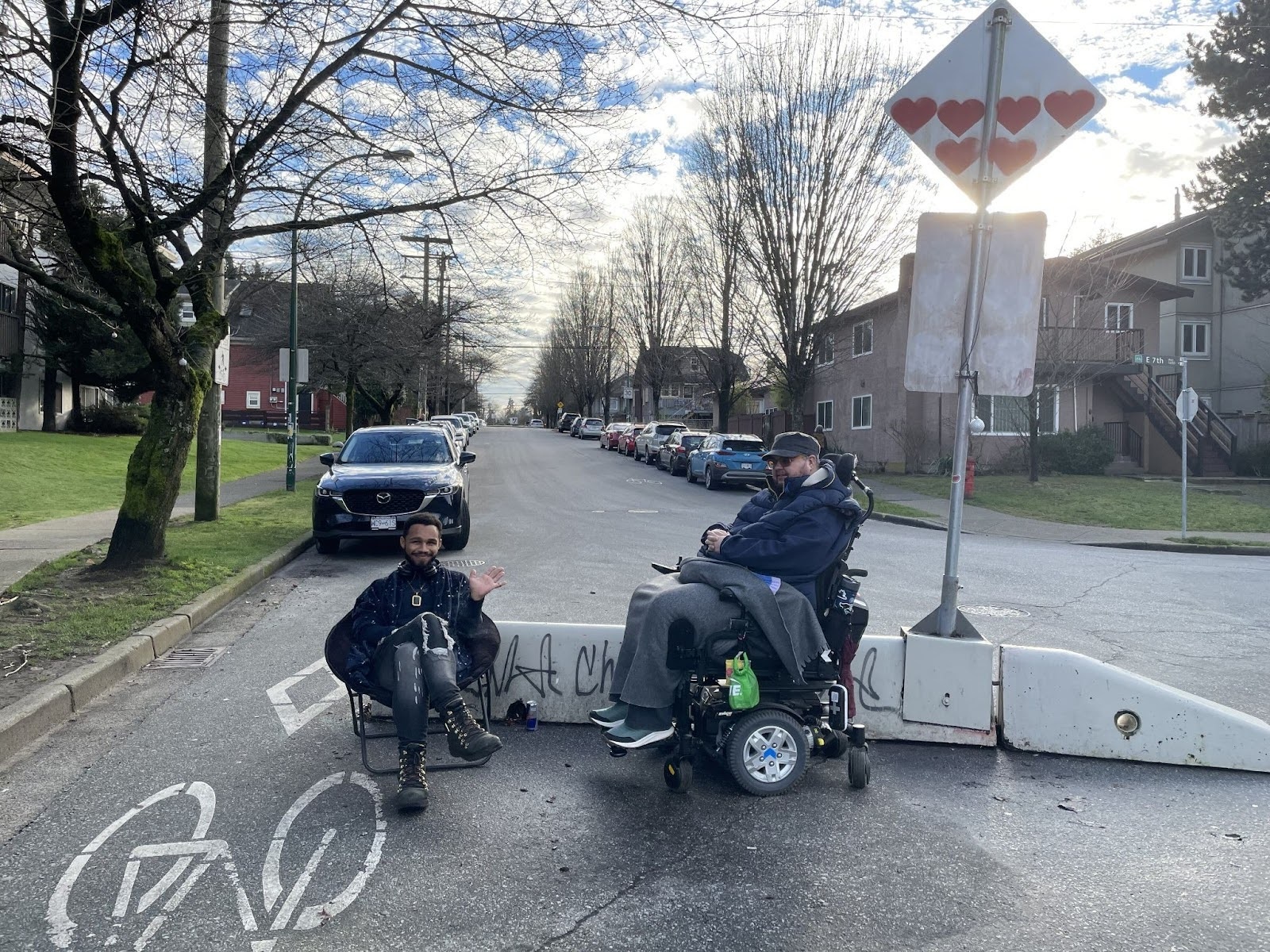 Two people in dark clothing are seated in front of a light grey concrete barrier in the middle of a residential street. Tajai, left, is waving and smiling at the camera. Logan, right, is in a motorized wheelchair and facing Tajai. It’s a clear day with fluffy white clouds in the sky.