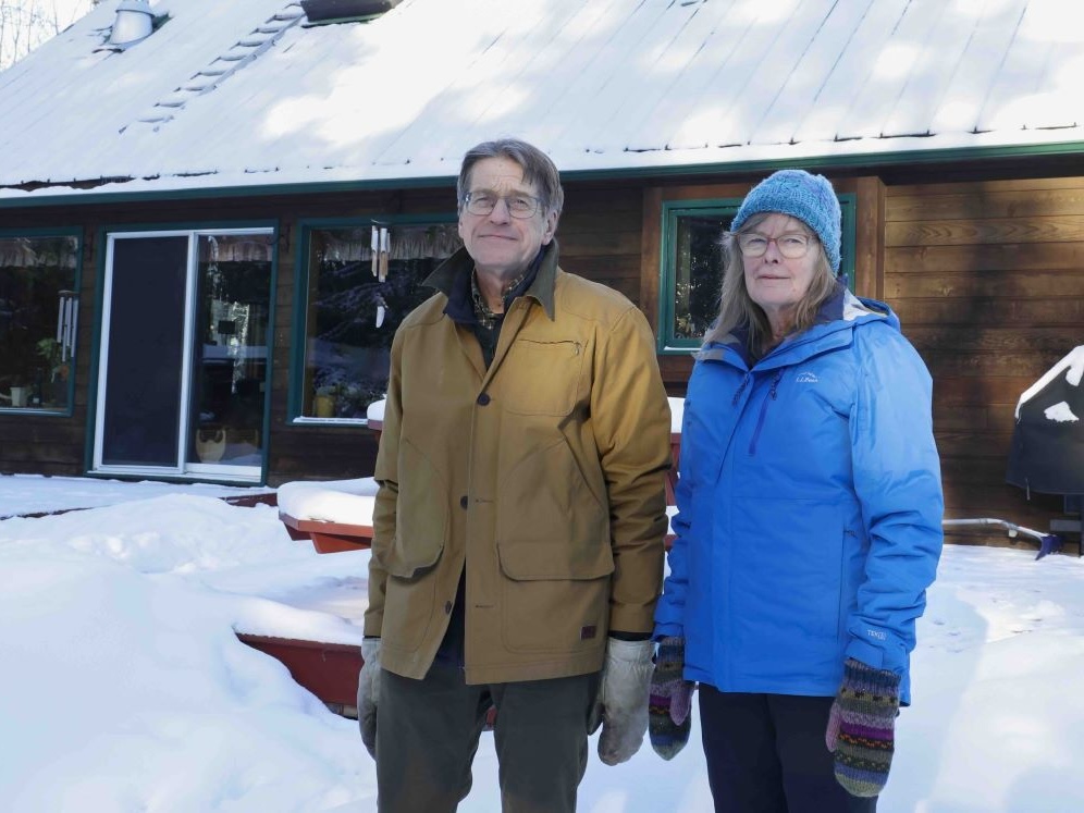 A woman and man wearing winter clothing stand in front of a snow-covered house in a rural setting.