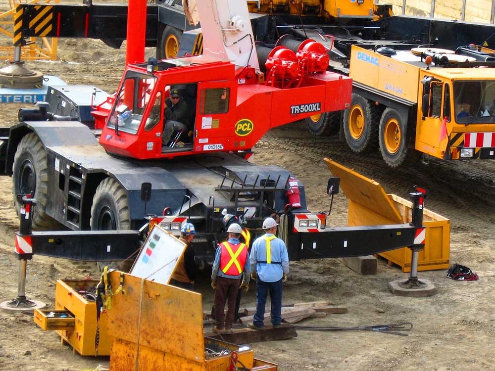 Two people in hard hats and visi vests stand on a construction site next to large cranes and machinery.