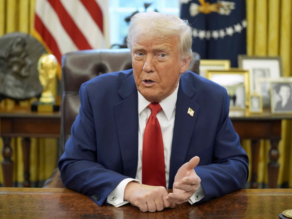 Donald Trump is seated at a desk in the Oval Office of the White House. He has dyed blond hair and tanned skin and is wearing a navy suit and red tie over a white shirt. Behind him is a U.S. flag and a desk full of framed photographs.