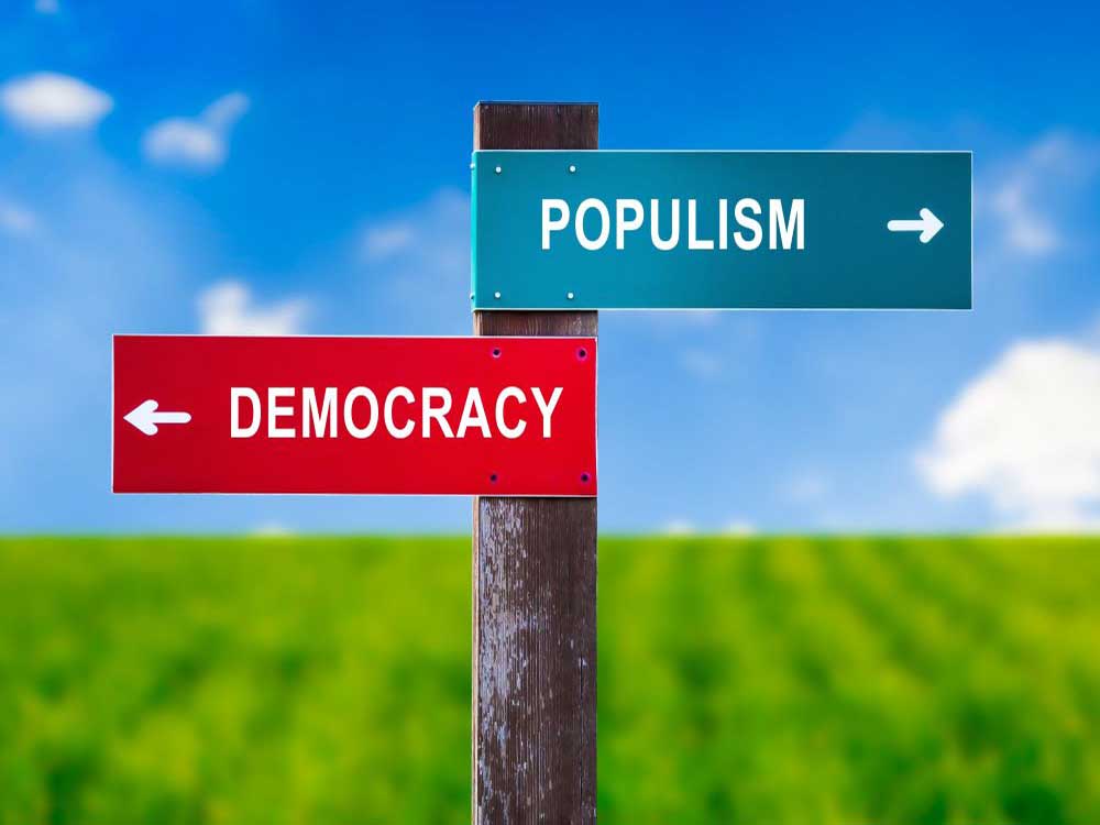 A signpost stands on a green pasture in front of a clear blue sky. A red sign reading 'Democracy' points to the left. A green sign reading 'Populism' points to the right.