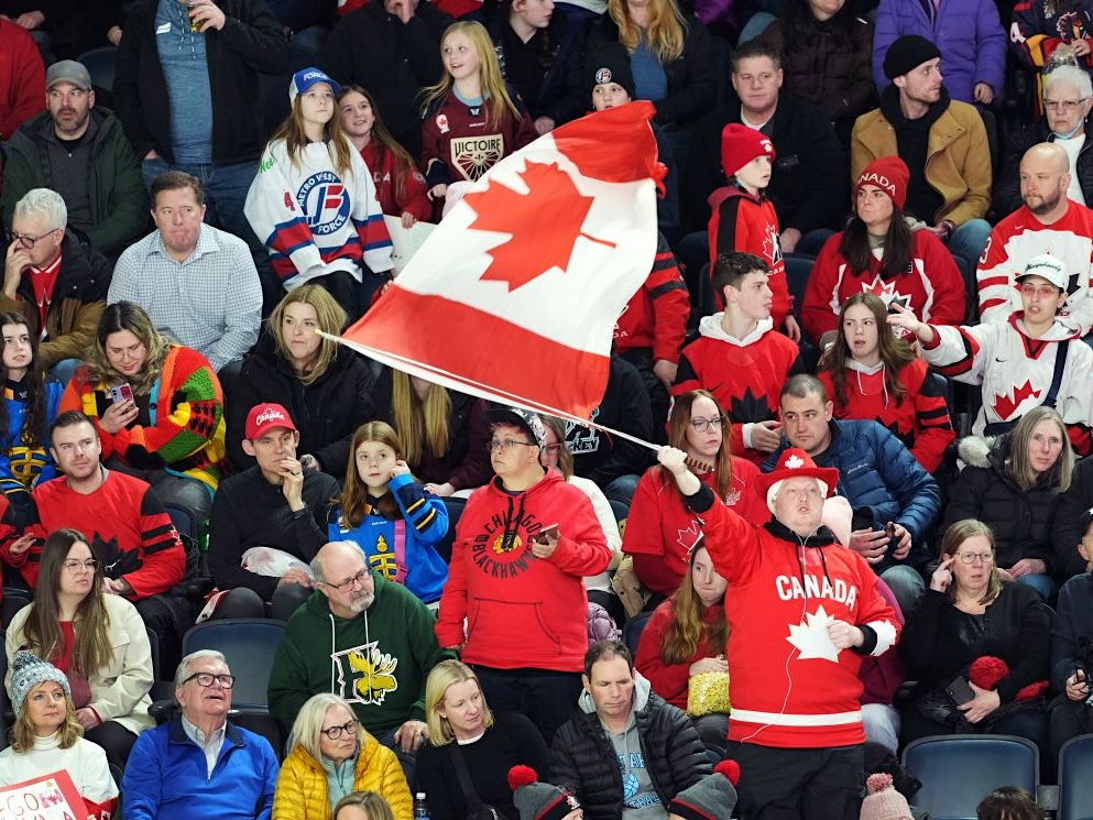 A man in a red Canada jersey and wearing a red cowboy hat with a white maple leaf on it waves a large Canadian flag. He is standing among rows of seated people, many dressed in red Canada-themed clothing.