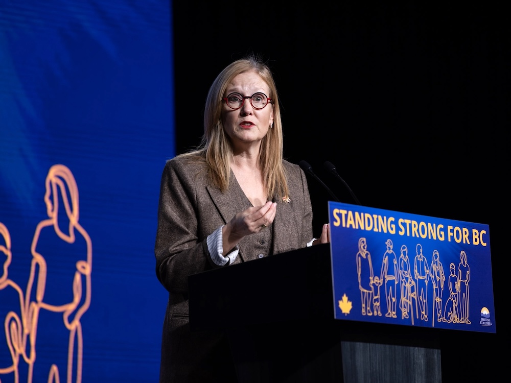 A 50-ish light-skinned woman with blond hair, wearing a brown suit, stands at a podium with a sign that says 'Standing Strong for BC.'