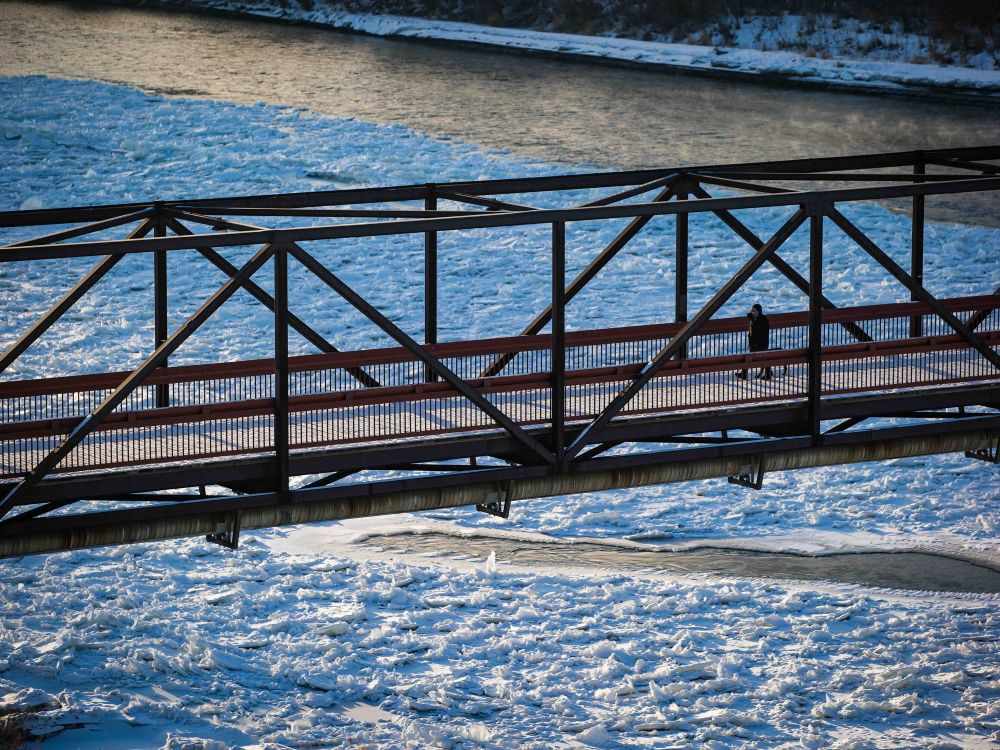 A pedestrian bridge with dark red beams spans across the snowy, icy North Saskatchewan River on a clear day in Edmonton.