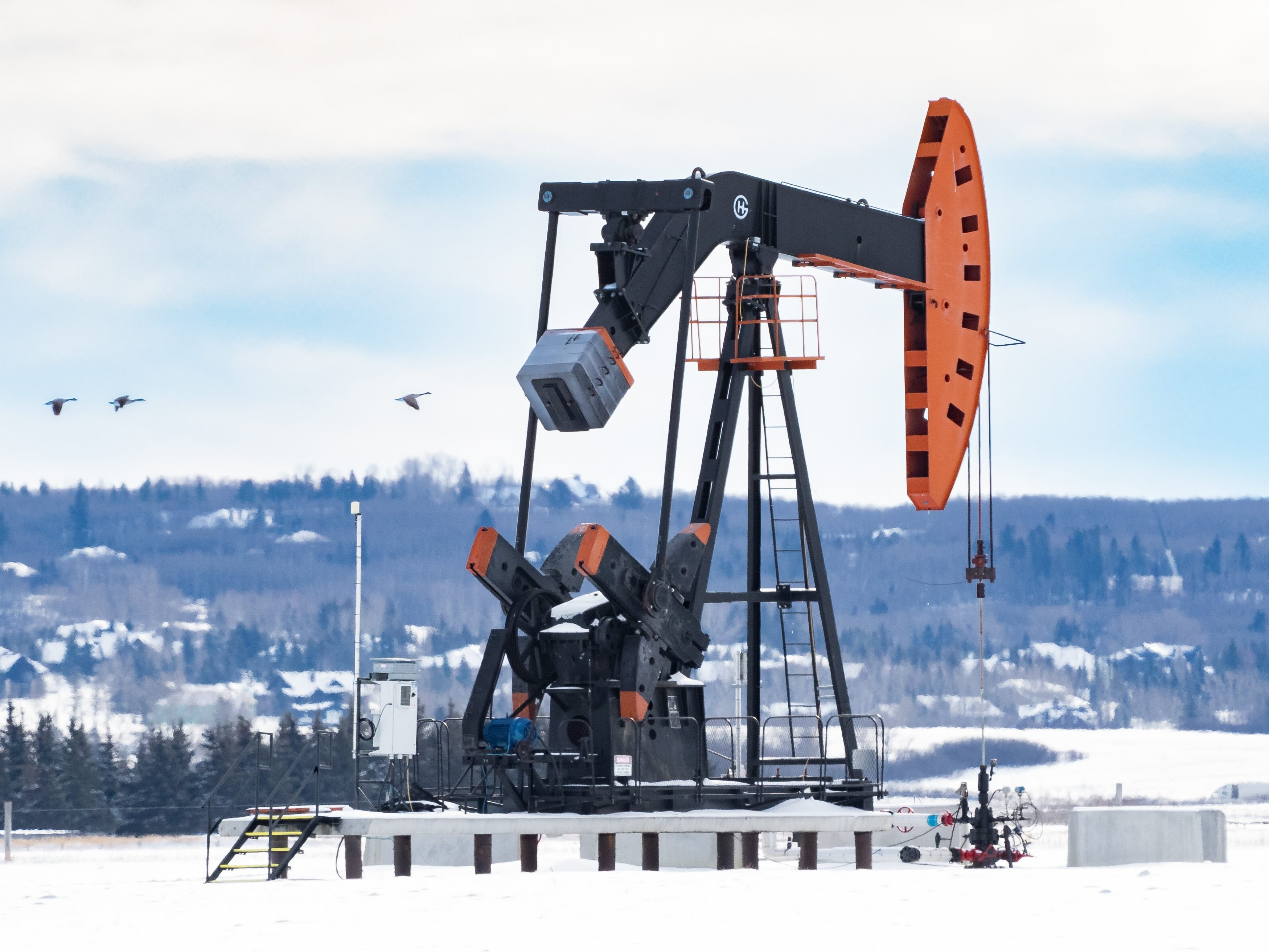 An oil well pumpjack stands in a snowy field, with a nearby community in the background.